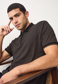 Bruin poloshirt met rits, voorzien van een getextureerd golfpatroon en zwarte accenten op de kraag en mouwen. Model leunt tegen een houten stoel.