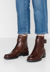 Bottes courtes en cuir marron avec un lacet sur le devant, une fermeture éclair latérale et une boucle décorative. Finition texturée et talon bloc bas.