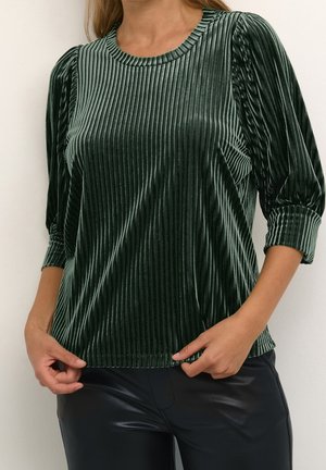 Frau trägt eine dunkelgrüne, samtige, gerippte Bluse mit dreiviertellangen Puffärmeln und schwarze Lederhose, die Hände in der Nähe des Saums.