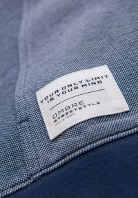Tissu bleu avec un tissage diagonal texturé. Une étiquette blanche affiche "VOTRE SEUL LIMITÉ EST VOTRE ESPRIT" et "OMBRE STREETSTYLE."