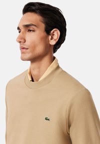 Sudadera beige con cuello alto y logo verde bordado en el pecho. Tela suave y lisa con un ajuste relajado y diseño minimalista.