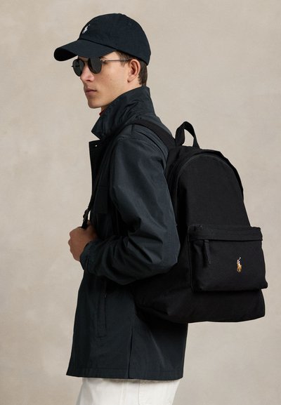 Hombre con gorra negra, gafas de sol, chaqueta oscura, pantalones blancos, llevando una mochila negra con un pequeño logo bordado en el bolsillo delantero.