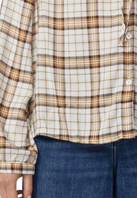 Kariertes Hemd in Braun- und Weißtönen, kombiniert mit blauen Jeans, zeigt den unteren Oberkörper und die linke Hand.