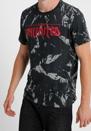 Mand iført sort og grå t-shirt med abstrakt mønster og rød tekst "UNLIMITED" samt sorte bukser, står med armene afslappet.