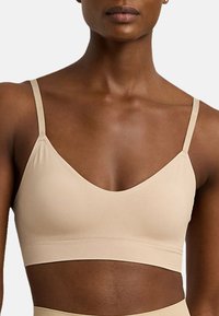Beige bralette med ett mjukt, stretchigt material. Har tunna, justerbara axelband och en djup V-halsringning. Slät yta, inga dekorationer.