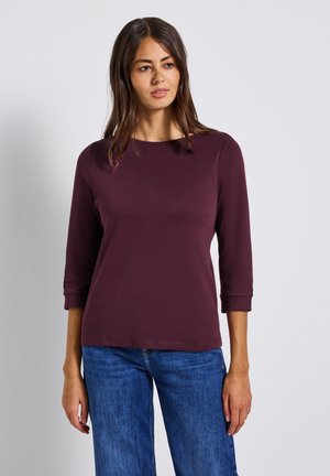 Vrouw met lang donker haar draagt een strakke paarse blouse met lange mouwen en een spijkerbroek, staande tegen een effen witte achtergrond.