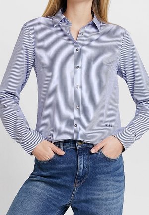 Femme portant une chemise boutonnée à rayures verticales bleues et blanches, rentrée dans un jean en denim bleu, les mains dans les poches.