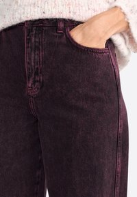 Jeans in denim bordeaux con una vestibilità classica, caratterizzati da cuciture visibili, una tasca laterale e una superficie del tessuto leggermente testurizzata.