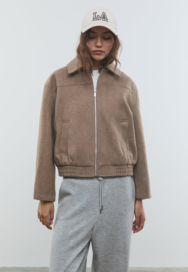 Veste bombardier en mélange de laine marron avec fermeture éclair à l'avant, poches latérales et poignets côtelés ; associée à un pantalon de survêtement gris à jambes larges et une casquette beige.