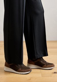 Brown sneaker featuring a mix of suede and mesh materials, with a textured upper, white sole, and contrasting accents. Paired with black pants.