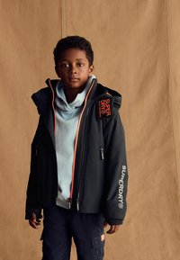 Giacca nera con dettagli arancioni e zip, indossata sopra un hoodie azzurro chiaro. Presenta il marchio "SUPERDRY" e tasche funzionali.