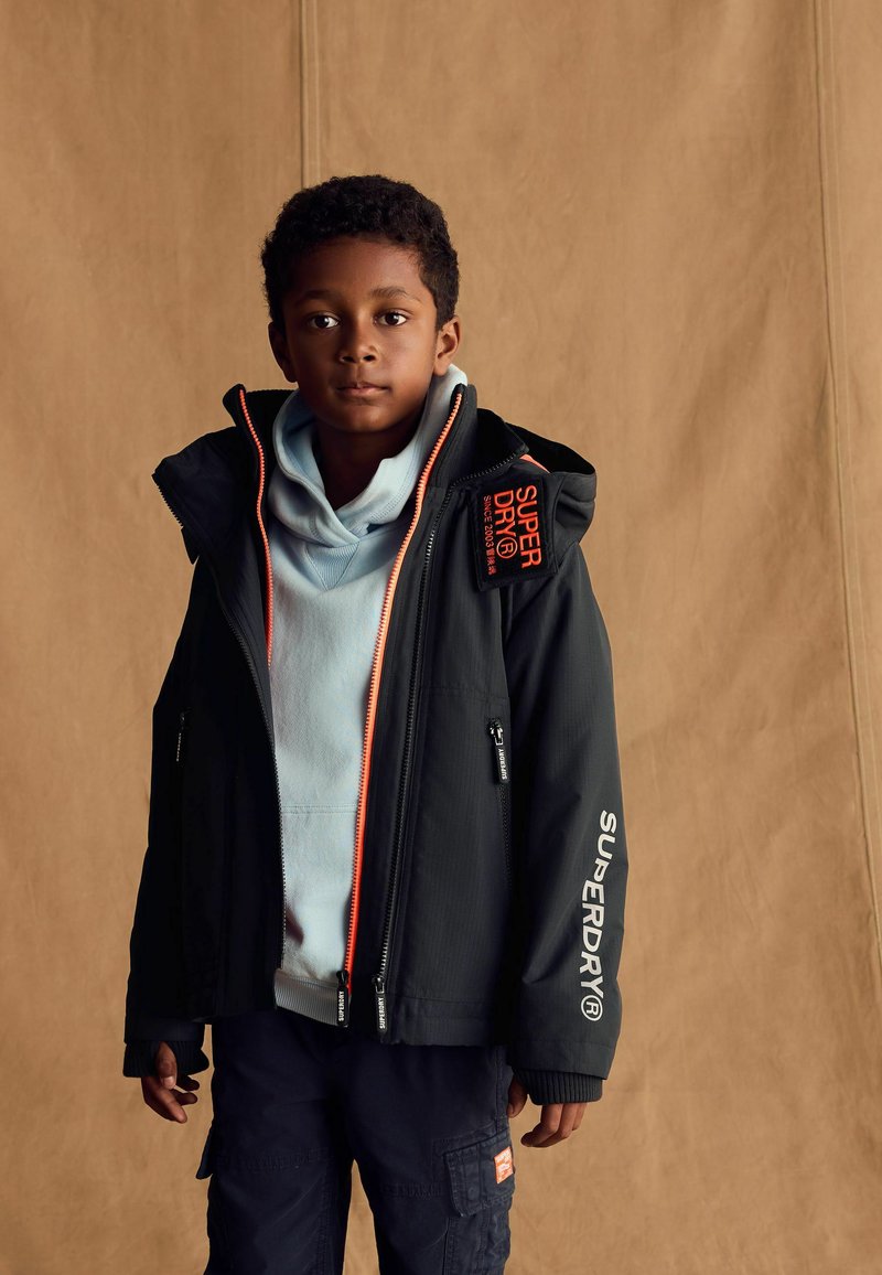 Giacca nera con dettagli arancioni e zip, indossata sopra un hoodie azzurro chiaro. Presenta il marchio "SUPERDRY" e tasche funzionali.