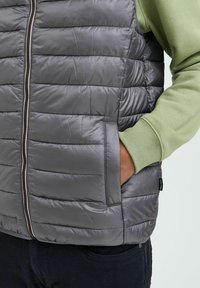 Chaleco de plumas acolchado gris con cremallera frontal, bolsillos laterales y textura suave. Lleva puesto un sudadera verde claro.