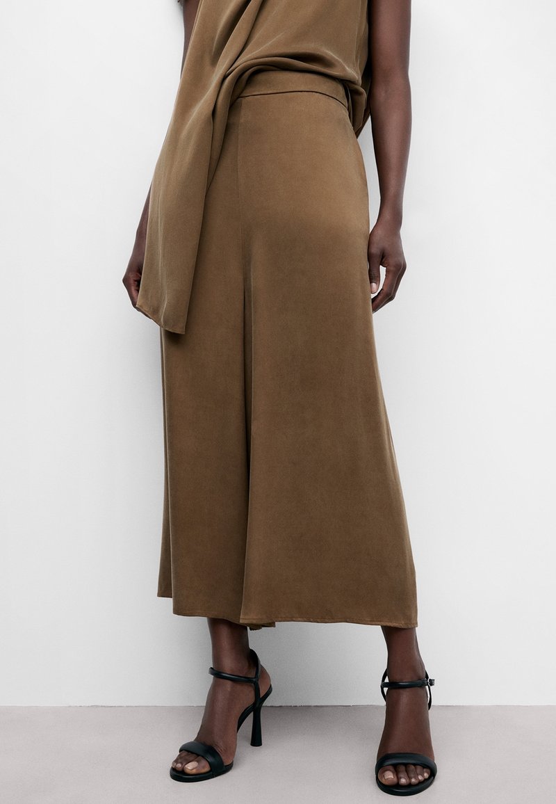 Femme portant un pantalon marron à jambes larges et des sandales noires à talons hauts, se tenant devant un fond blanc uni.