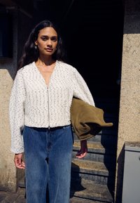 Hvid tekstureret cropped jakke med lynlås, kombineret med højttaljede blå denimjeans. Holder en brun ruskinds taske. Lys udendørs omgivelser.