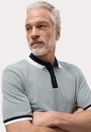 Camiseta polo gris con un diseño texturizado, cuello y botones en color marino, y ribete blanco en las mangas. El modelo tiene los brazos cruzados.