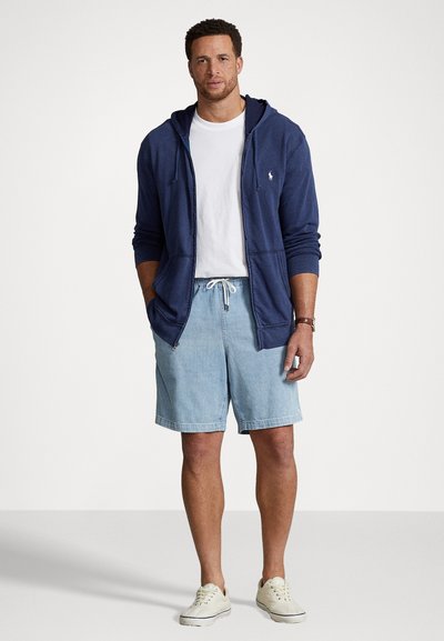 Morska zip-up dukserica preko bijele majice, uparena s svijetloplavim traper kratkim hlačama. Bijele tenisice upotpunjuju ležerni outfit.