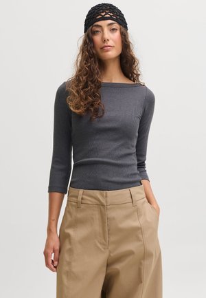 Femme aux longs cheveux bouclés portant un bandeau noir en résille, un haut gris foncé à manches longues et un pantalon beige taille haute, les mains dans les poches.