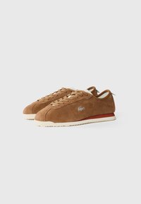 Brown suede sneakers with a low profile, white rubber sole, and beige laces. Features a silver logo on the side and a red accent on the heel.