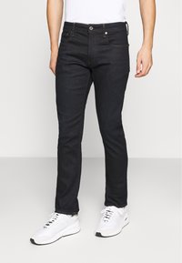 Jeans de mezclilla azul oscuro con corte ajustado, con costuras naranjas, cierre de botón y bolsillos delanteros y traseros. Combinados con zapatillas blancas.