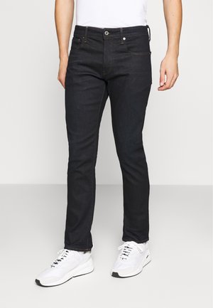Jeans de mezclilla azul oscuro con corte ajustado, con costuras naranjas, cierre de botón y bolsillos delanteros y traseros. Combinados con zapatillas blancas.
