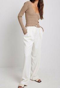 Cardigan marrone a coste con scollo a V e chiusura con bottoni, abbinato a pantaloni ampi plissettati bianchi e sandali neri a scivolo.