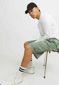 Modèle assis sur une chaise, portant une chemise blanche à manches longues et un short cargo vert olive. Les accessoires incluent une casquette noire et des baskets blanches avec des rayures noires.