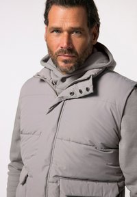 Graue gepolsterte Weste mit hohem Kragen, die mit Druckknöpfen und zwei Fronttaschen ausgestattet ist, über einem grauen Hoodie getragen. Glatte Textur und modernes Design.