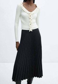 Top blanco de manga larga con textura acanalada y cuello con volantes, combinado con una falda negra plisada. La falda presenta líneas verticales y llega hasta los tobillos.