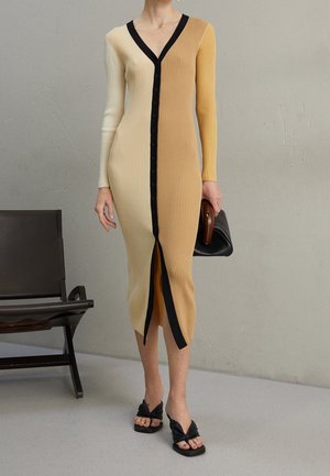 Woman wearing a ribbed, two-tone beige and mustard button-up midi dress with black trim, holding a black clutch and wearing black sandals.