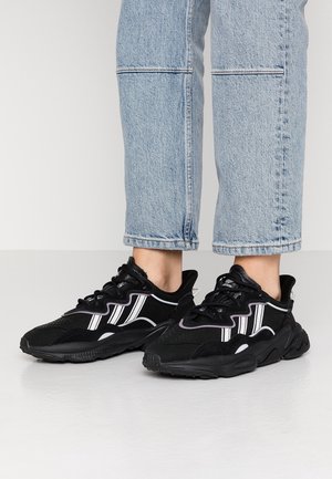 Person iført lyseblå korte jeans og sorte sporty sneakers med hvide striber, der står på en grå gulv.