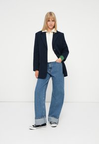 Donna bionda in piedi contro uno sfondo bianco, che indossa un blazer blu navy, un maglione bianco con zip, jeans blu a gamba larga con risvolto e sneakers nere.