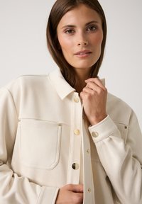 Frau mit glattem braunen Haar, die eine cremefarbene Jacke mit Knöpfen und zwei Brusttaschen trägt und den Kragen nah an ihrem Hals hält.
