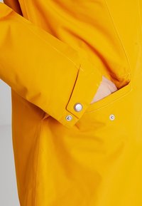 Veste imperméable jaune avec un tissu texturé, des boutons-pression et une poche latérale intégrée au design.