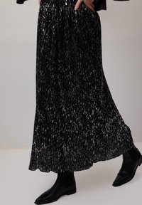 Long black pleated skirt with silver speckles worn with black ankle boots and a hand resting in pocket.