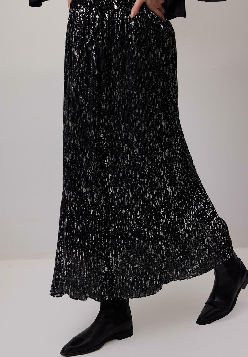 Long black pleated skirt with silver speckles worn with black ankle boots and a hand resting in pocket.