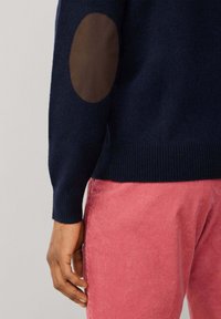 Marineblauer Pullover mit einem braunen Ellbogenpatch, gerippten Manschetten und einem gerippten Saum, kombiniert mit rosafarbener Cordhose. Fokus auf Texturunterschiede und Farben.
