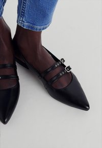 Schwarze Lederballerinas mit spitzen Zehen, ausgestattet mit zwei dünnen Riemen, silbernen Hardware-Elementen und einem niedrigen Blockabsatz.