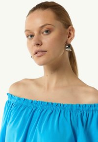 Niebieska bluzka bez ramiączek z falbaną przy dekolcie; zestawiona z dużymi metalicznymi kolczykami. Gładki materiał i błyszczący, okrągły kształt kolczyków.