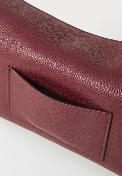 Burgundy läderväska från sidan med en texturerad yta och en liten extern ficka. Formad med rundade kanter som framhäver materialets kvalitet.