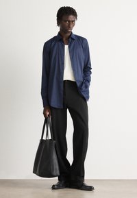 Camisa de botones azul marino, camisa interior blanca, pantalones negros y bolso de cuero negro. Colores sólidos, diseño depurado y estética minimalista.