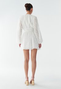 Vit spetsklänning med långa, luftiga ärmar, markerad midja och utsvängd kjol. Har ett texturerat mönster och ett lätt, genomskinligt tyg.