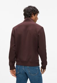 Barna kapucnis pulóver, texturált felülettel, álló gallérral és bordázott mandzsettákkal, kék farmernadrággal párosítva. Hátsó nézet látható.