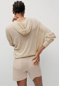 Sudadera beige de punto con capucha, ajuste holgado y pantalones cortos con bolsillo trasero. Cuenta con capucha y puños de canalé, textura suave y diseño casual.