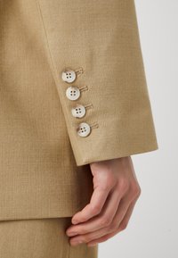 Sako v béžové barvě s rukávem zdobeným čtyřmi kulatými, světle zbarvenými knoflíky se švy, které ukazují texturovanou tkaninu.