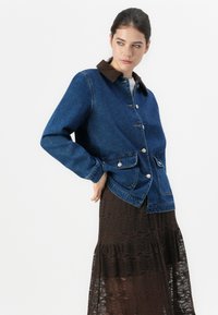 Kvinne iført en blå denimjakke med knapper foran og klaffelommer, kombinert med et mørkebrunt blondeskjørt, ser bortover.