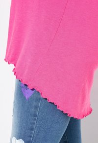 Rosa T-Shirt mit gewelltem Saum und kurzen Ärmeln, aus einem weichen, leichten Stoff, kombiniert mit verwaschenen blauen Jeans, die einen lila Patch aufweisen.