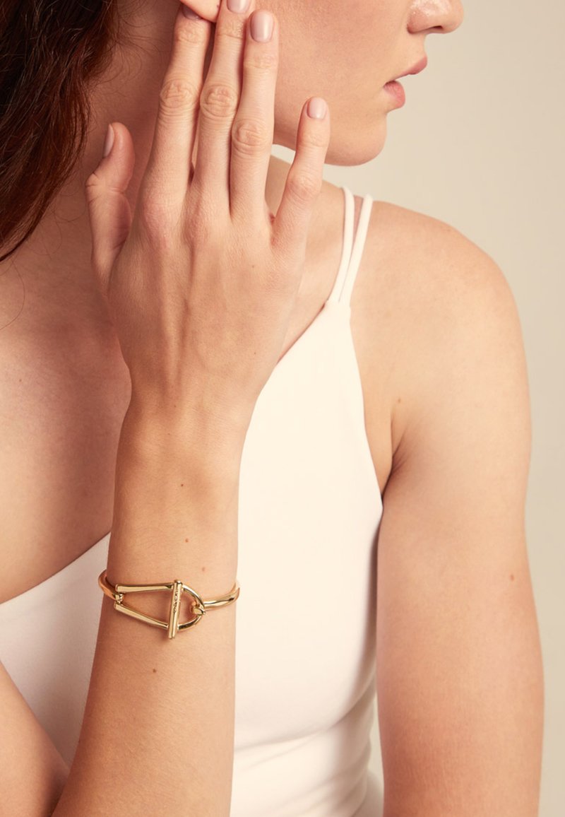 Pulsera de brazalete de oro con un diseño geométrico simple que presenta un círculo abierto y un acento de barra recta. Textura suave y pulida.