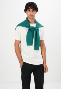 Ung mand med mørkt, krøllet hår iført hvid t-shirt med logo, sorte bukser, og en blågrøn sweater draperet over skuldrene, med den ene hånd i lommen.
