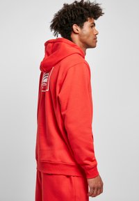 Red hoodie with front pocket and adjustable drawstring. Features a logo on the back, made from a soft, textured fabric.